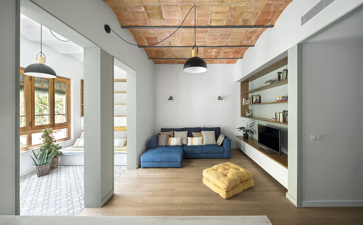 Salón con sofá azul y puf amarillo, mueble televisión en hueco pared techo con bóveda catalana