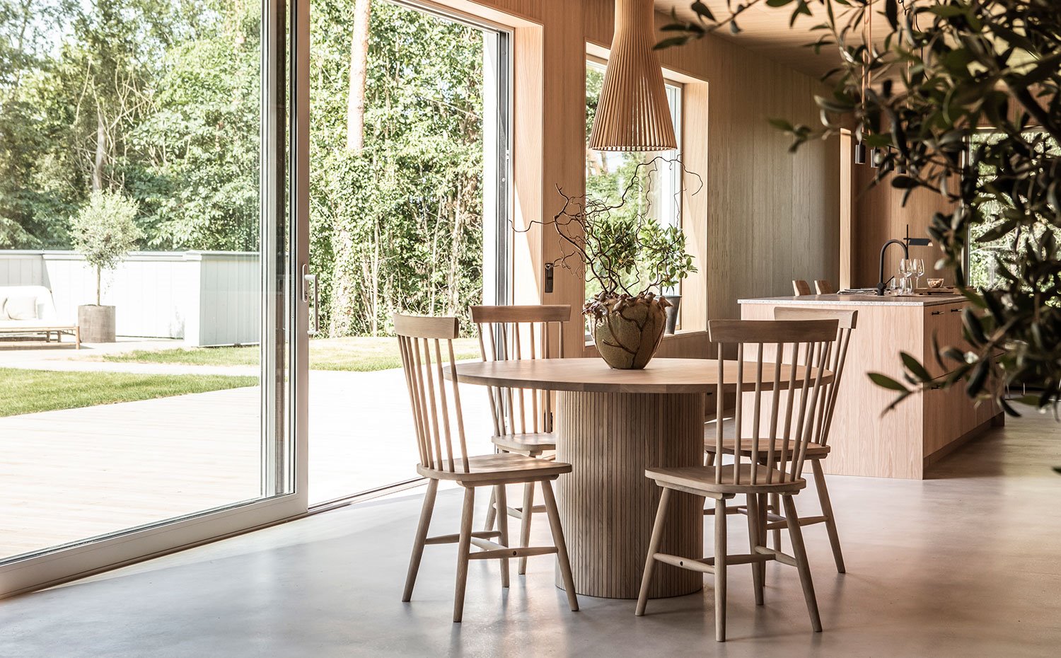 Comedor con mesa circular, sillas y luminaria suspendida, todo de madera, con grandes aperturas hacia el exterior