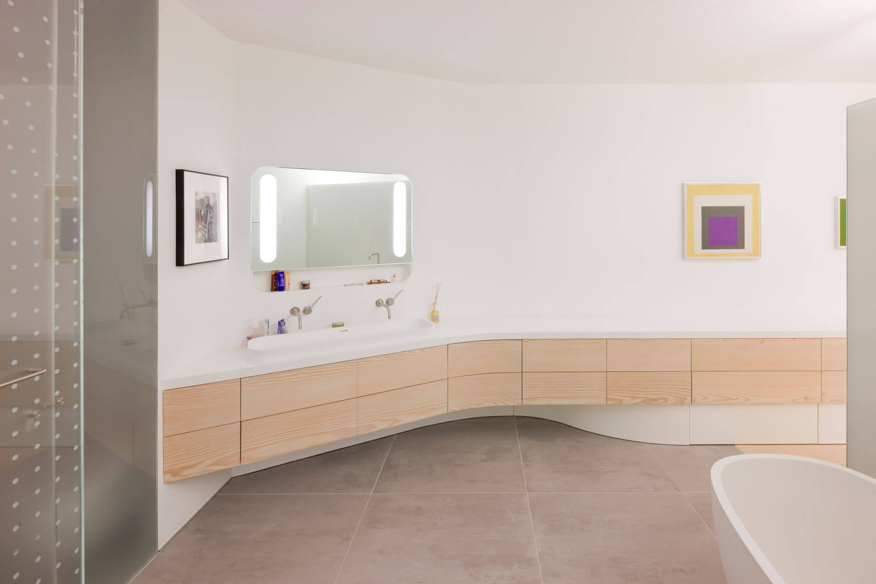 10. Presidido asimismo por obras de arte, el baño es alargado y expresa simplicidad. El lavabo se integra en una encimera serpenteante sobre un mueble suspendido con frentes de madera y sin tirador. La bañera es de Boffi