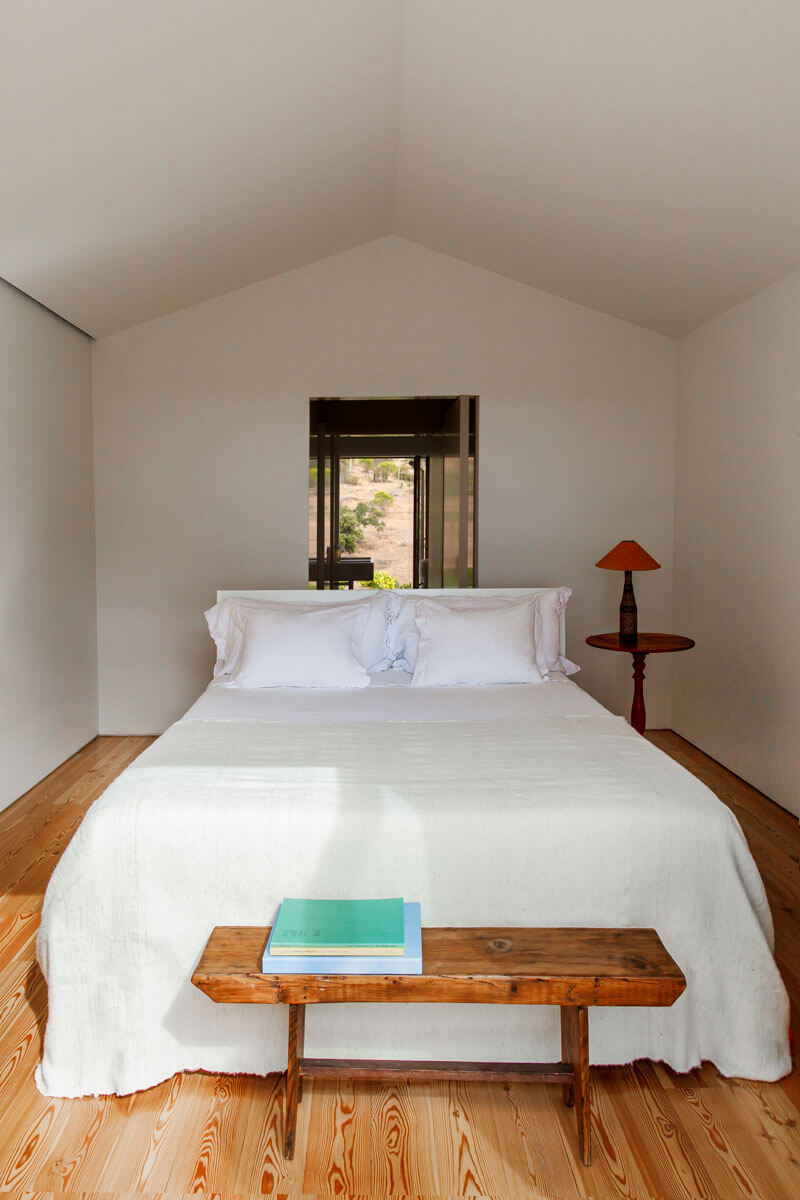 7b-. La cama de matrimonio preside el dormitorio, un espacio escueto que tiene como única decoración un banco y una mesita de madera. El pavimento de madera de pino y los armarios empotrados a los lados de color blanco construyen la piel de esta habitación
