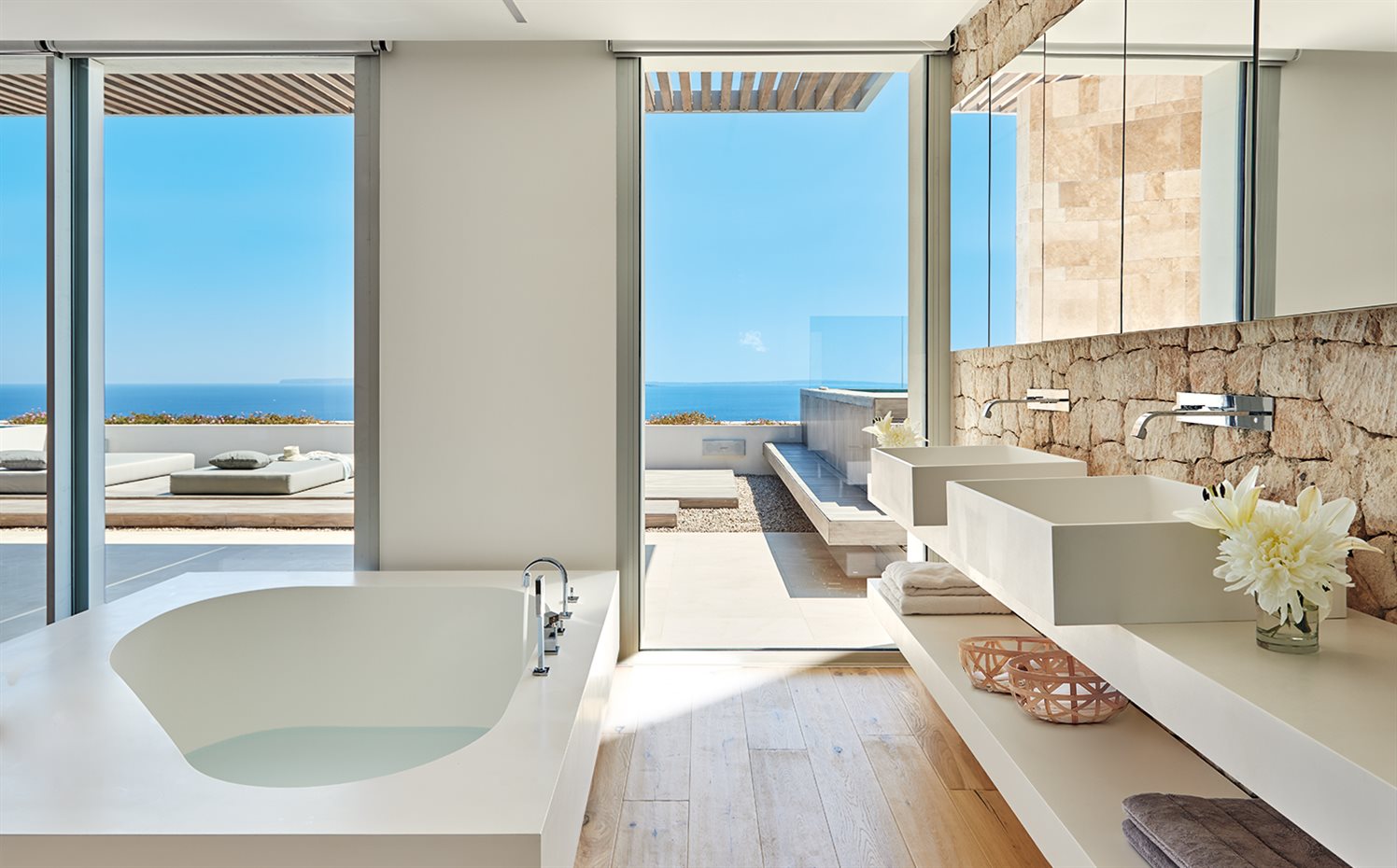AyD-Saota(Ibiza)2252b. La pared de piedra del baño proporciona un fondo de calma sólida a esta estancia abierta a la terraza. Una solidez atemperada por la ligereza de los tonos claros del suelo de madera y de los sanitarios