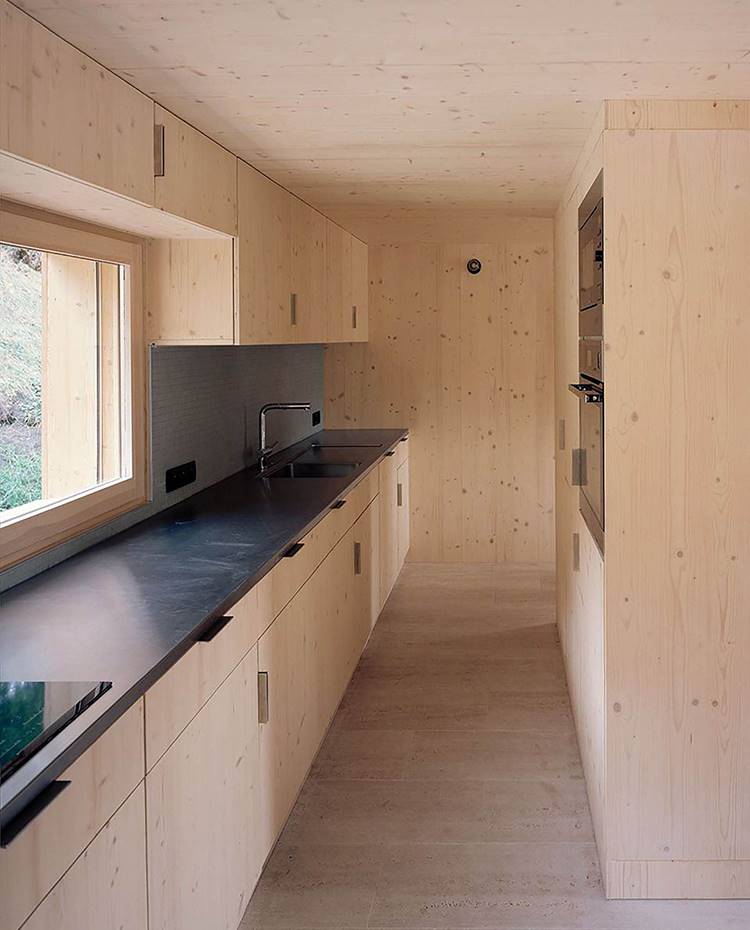 cocina en línea, mobilario de madera, electrodomésticos y encimera en negro, ventana sobre encimera. Para la cocina, Joachim Fritschy proyecta una distribución en línea, totalmente revestida con la misma madera que el resto de la vivienda y cuya (omni)presencia solo es interrumpida por los electrodomésticos y la encimera en negro.