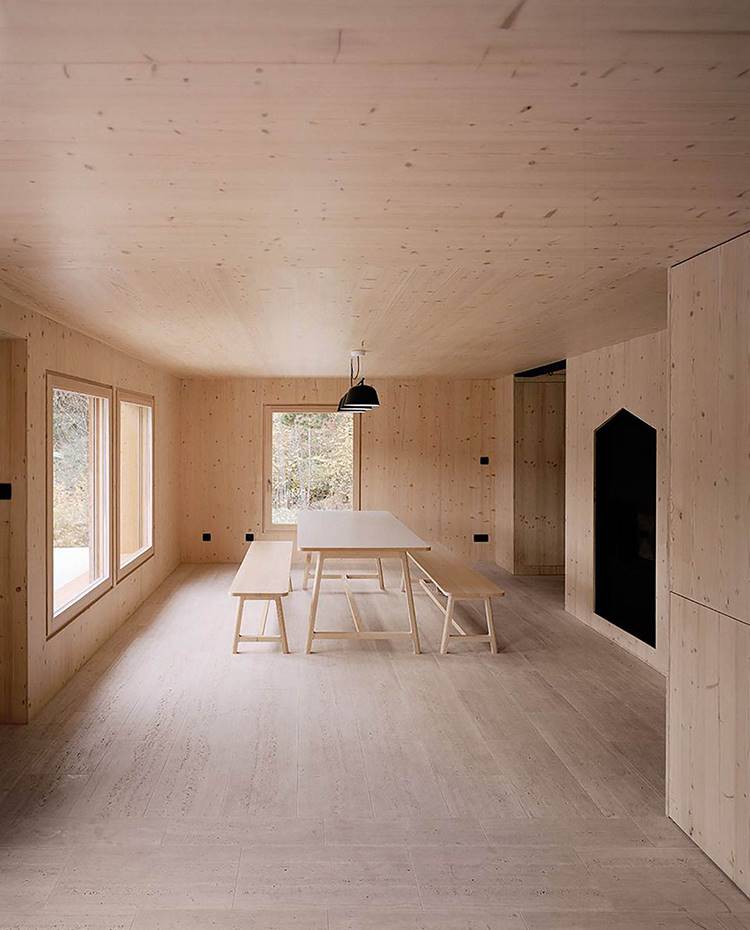 comedor revestido de madera clara, mesa de madera, bancadas de madera, lámparas de techo negras, ventanas al exterior alpino. El comedor se viste con piezas de madera que se mimetizan con el espacio. La presencia de grandes ventanales beneficia la entrada de luz natural y a su vez convierte a la estancia en un escaparate que se proyecta hacia el paisaje exterior.