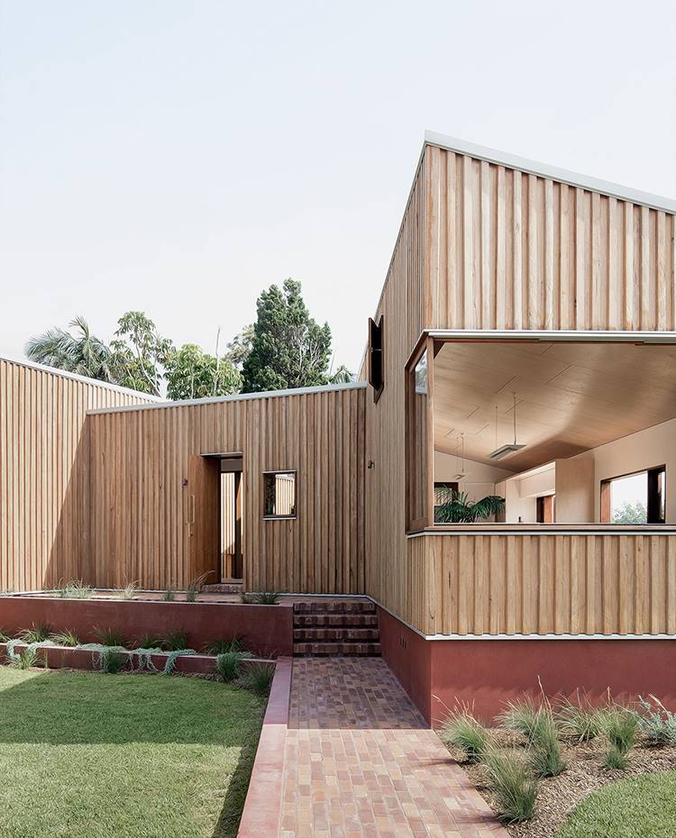 Entrada principal vivienda, exterior, frente ajardinado. Una fachada alistonada de madera y grandes ventanales que se extienden hacia el exterior, con repisas para tomar asiento en la propia apertura, dan forma al capazarón de la vivienda.