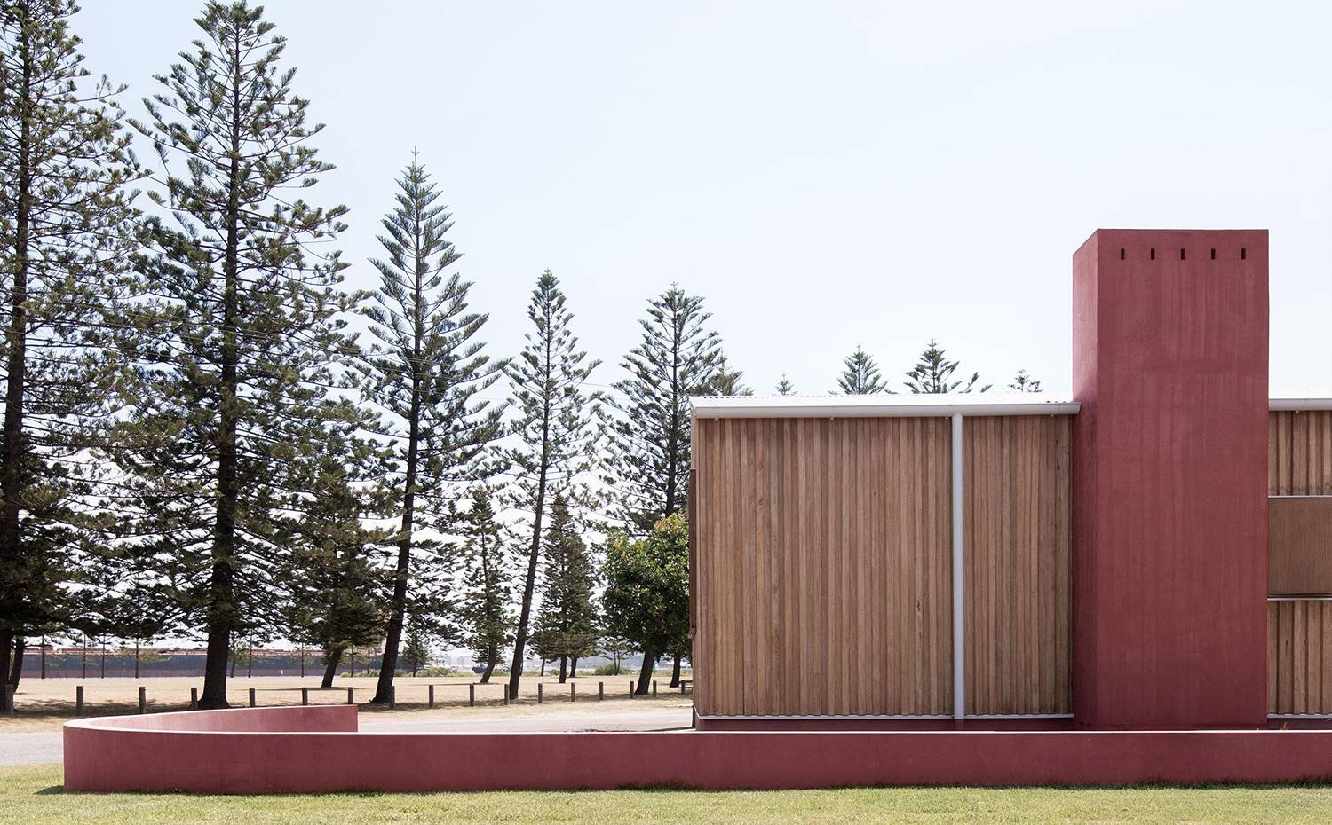 Exterior vivienda, árboles, pequeño murete alrededor, revestimiento madera. Un bajo muro de color rojo tierra se curva alrededor de la casa delimitando el terreno. Dicho color, también presente en la fachada, alude al rojo oxidado de los barcos cargados de carbón que circulan por el puerto de Newcastle. 