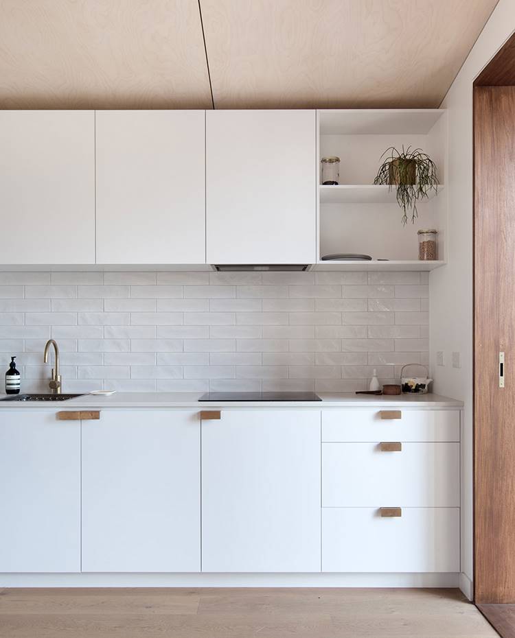 Frontal cocina blanca, campana extractora integrada, frontal baldosa rectangular, suelo madera. La cocina se integra en el módulo de dia de una forma discreta, con un mobiliario lacado en blanco y frontal cerámico del mismo color. 