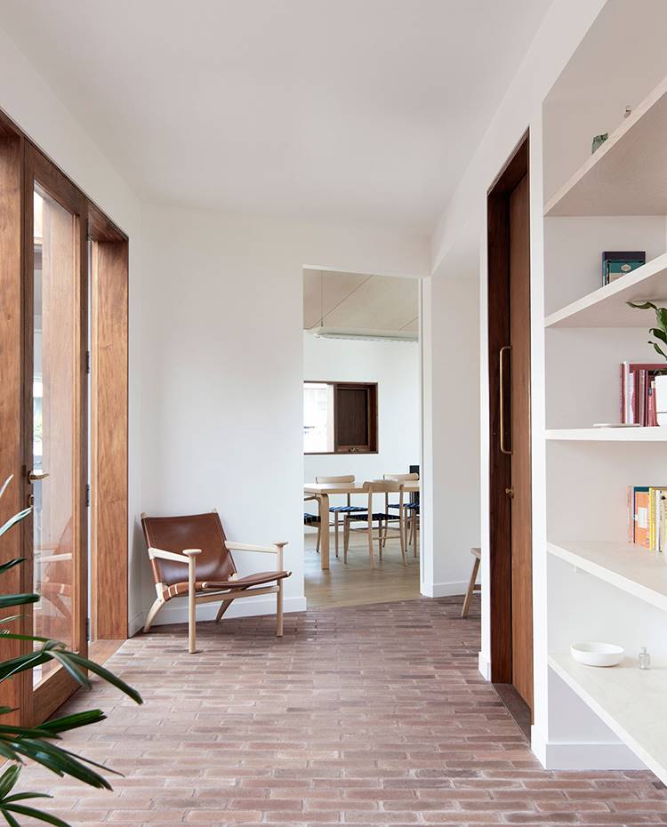 Zona de paso, estantería junto a pared, butaca de piel y madera, comedor de fondo . Luminosa y expansiva, la zona de lectura es a la vez el puente entre el módulo de día y el de noche. Una zona diáfana y serena gracias a los cerramientos acristalados y un mobiliario sencillo y funcional, como las estánterías hechas de obra.