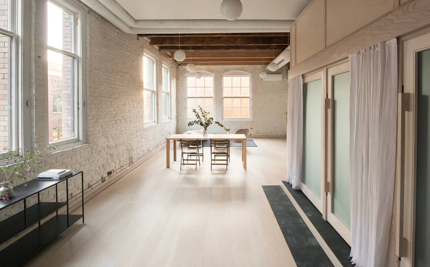 Consola negra, suelo de madera, lof inustrial, pared de obra vista, puertas de cristal esmerilado. Junto a la entrada de luz natural que incide por las ventanas, la luz artificial procede de luces colgantes blancas en forma de globo, distribuidas por todo el apartamento.
