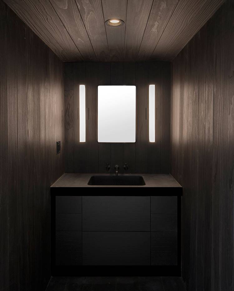 Lavamanos baño, revestimiento de madera, ambiente oscuro, suelo baldosa de cemento. Interior del espacio destinado a tocador, con lavamanos de piedra, paredes revestidas de madera oscura y suelos cubiertos de azulejos de cemento oscuro, para contrastar los tonos pálidos del resto de la casa.