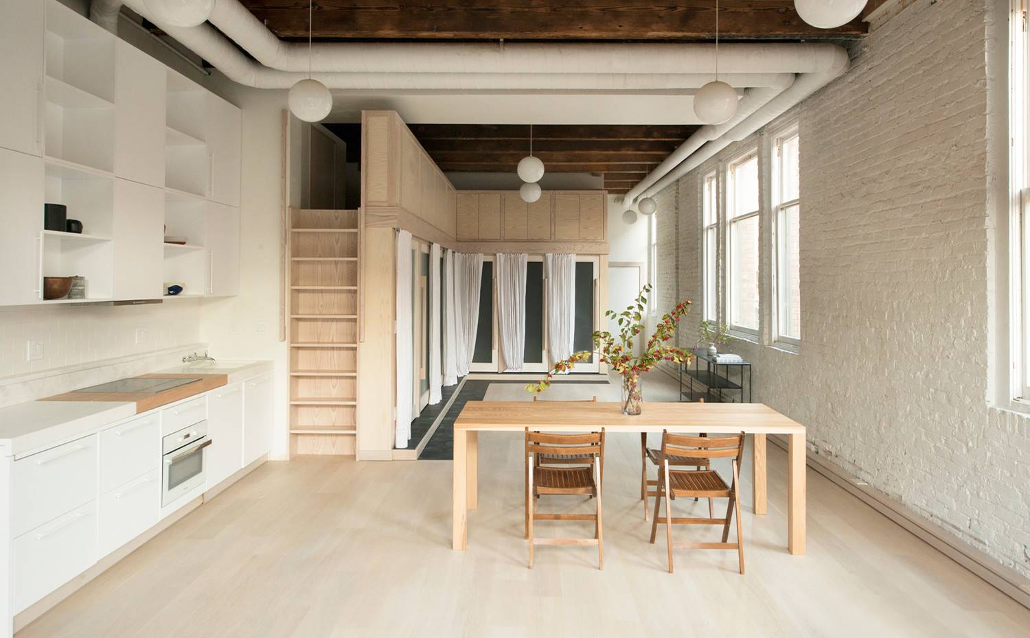 Mesa comedor, sillas plegables madera, escalera vertical, cocina blanca, conductos de aire en blanco, loft industrial. La mesa de comedor hecha a medida, en madera de nogal, es de los pocos muebles que visten el apartamento. Se acompaña de sillas plegables de madera y una luminaria de suspensión blancas, tipo globo, que contrasta con las vigas y los paneles de madera oscura del techo.