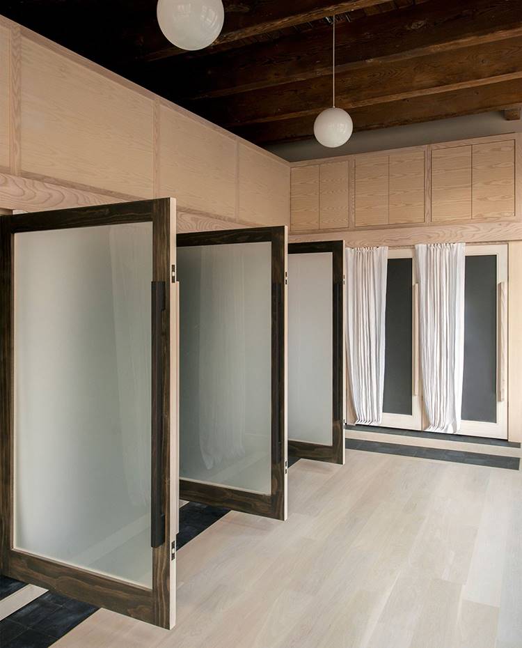 Puerta abiertas en cristal esmerilado, baño en loft, suelo de madera, estructura de madera, lámparas suspendidas tipo globo. Con el objetivo de sobredimensionar la entrada en cada uno de los espacios que conforman la zona de baño, el equipo de interioristas de Le Whit ha incluido un escalón y enormes tiradores de roble al frente de cada una de las puertas.