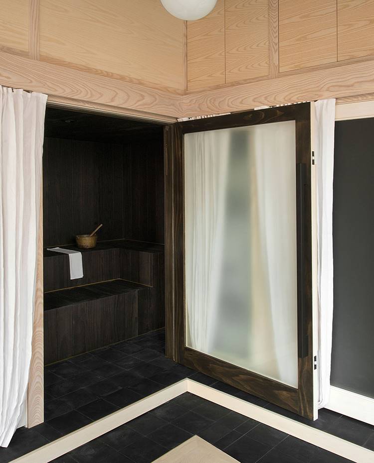 Sauna con puerta abierta, revestida de madera y suelo de baldosas de cemento negras, puerta en cristal esmerilado, lámpara suspendida tipo globo. Las puertas de vidrio esmerilado brindan acceso a cada una de las unidades, que están forradas en el interior con madera calcinada usando la técnica tradicional japonesa Shou Sugi Ban, que hace que el material sea resistente a la podredumbre y al fuego. 