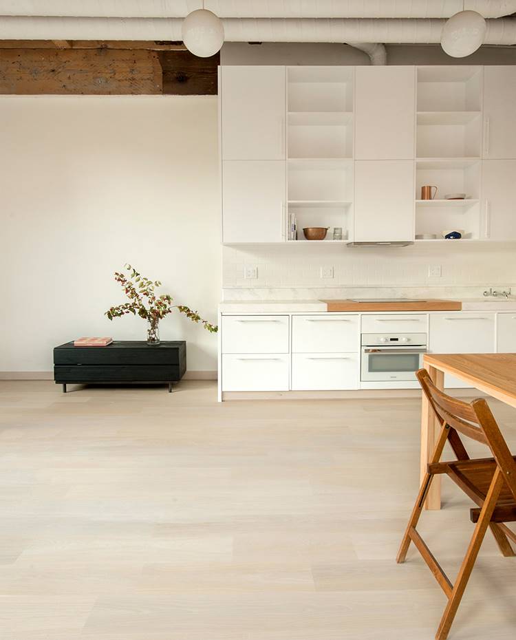 Cocina blanca en línea, mesa de madera, silla plegable, módulo auxiliar negro, conductos de aire a vista. Cocina abierta al espacio diáfano e industrial del apartamento, cuya encimera de mármol brillante integra una tabla de madera de nogal en la zona de trabajo. La gran altura de la vivienda permite gran capacidad de almacenamiento vertical.