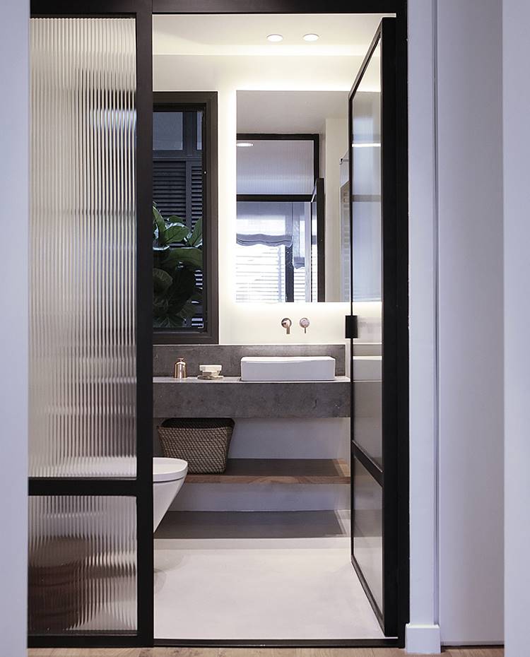 Cerramiento baño cristal esmerilado, sanitario suspendido, lavamanos sobre encimera piedra. En los baños, todo se apoya en los materiales utilizados: la piedra San Vicente, el microcemento beige y el cobre acaparan todo el protagonismo. Eso sin hablar del cerramiento en cristal esmerilado que deja pasar la luz procedente del exterior, preservando la intimidad de la estancia.