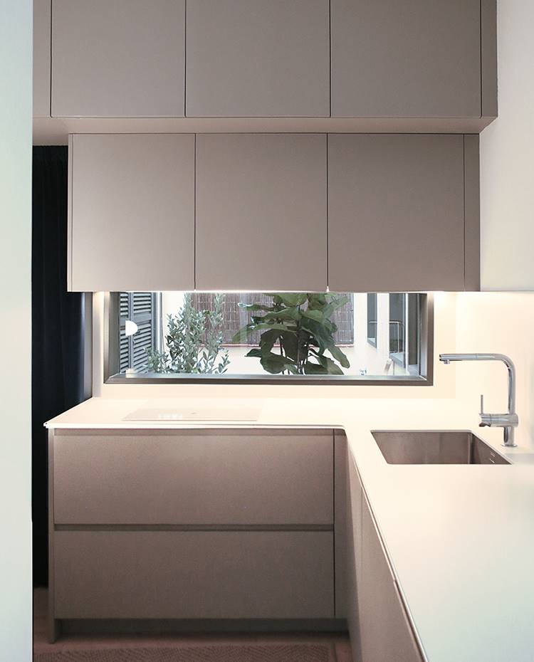 Cocina en forma de L, zona de aguas y cocción, dobme nivel mobiliario superior. La cocina en forma de L se vuelca sobre un patio privativo que le brinda de claridad y buena ventilación. El mobiliario, en línea con el resto de la vivienda, se tiñe de tonos grises y beiges.