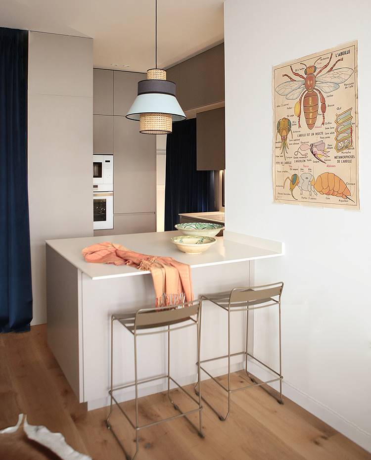 Cocina semi-integrada comedor, taburetes de office en península, lámpara suspendida. La cocina está semintegrada al salón-comedor a través de una península que hace las veces de zona de 'office', acompañada de dos taburetes de suaves líneas. El diseño de la lámpara de suspensión juega con la rejilla vegetal, material omnipresente en el proyecto.