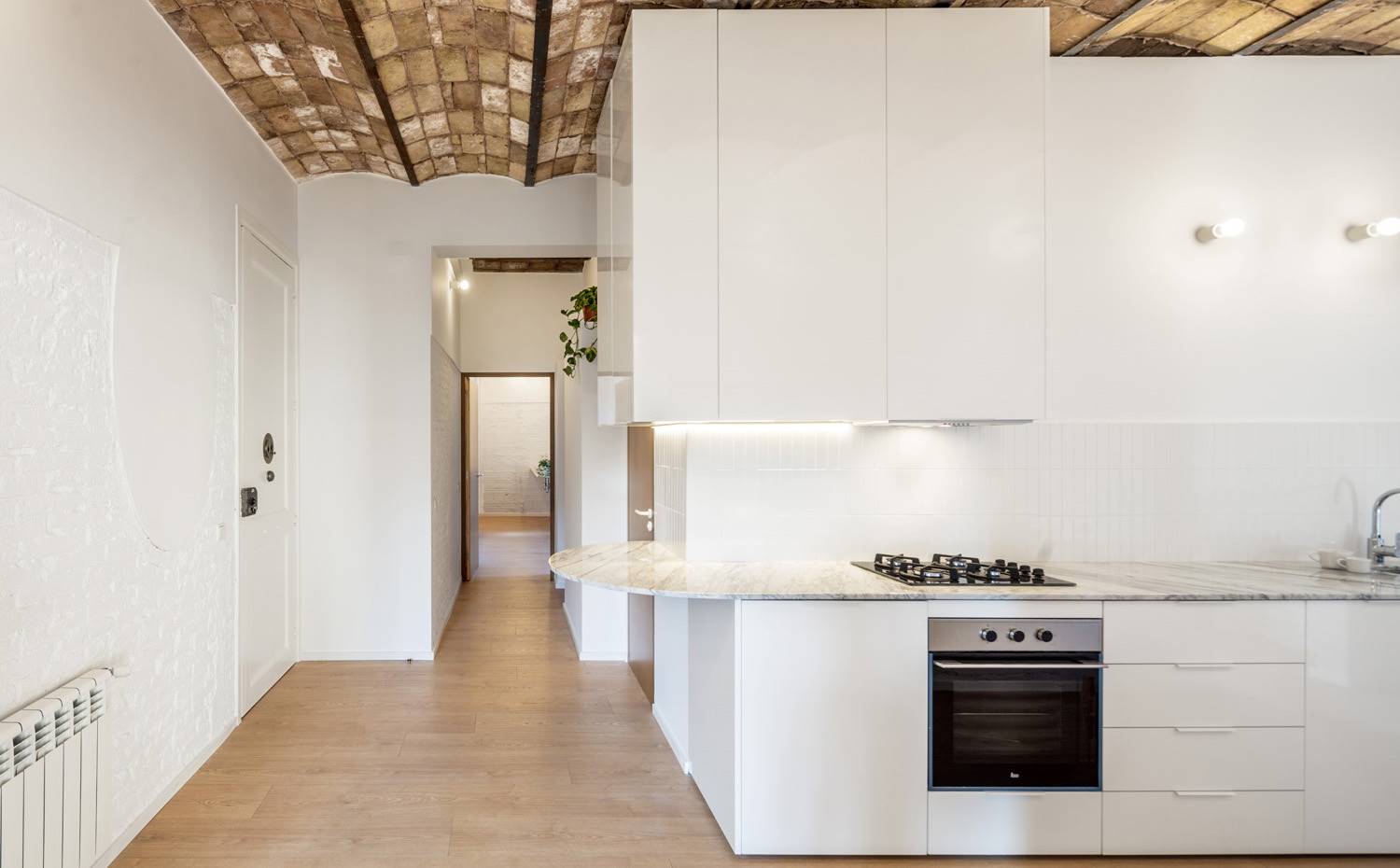 Cocina blanca, abierta, techo volta catalana, suelo de madera. El proyecto cuenta con dos planos claramente definidos. Por un lado está el horizontal, que aporta calidez a base de suelos de madera y techos de bóveda catalana; y por el otro las superficies verticales, que brindan neutralidad a base de acabados en blanco.   