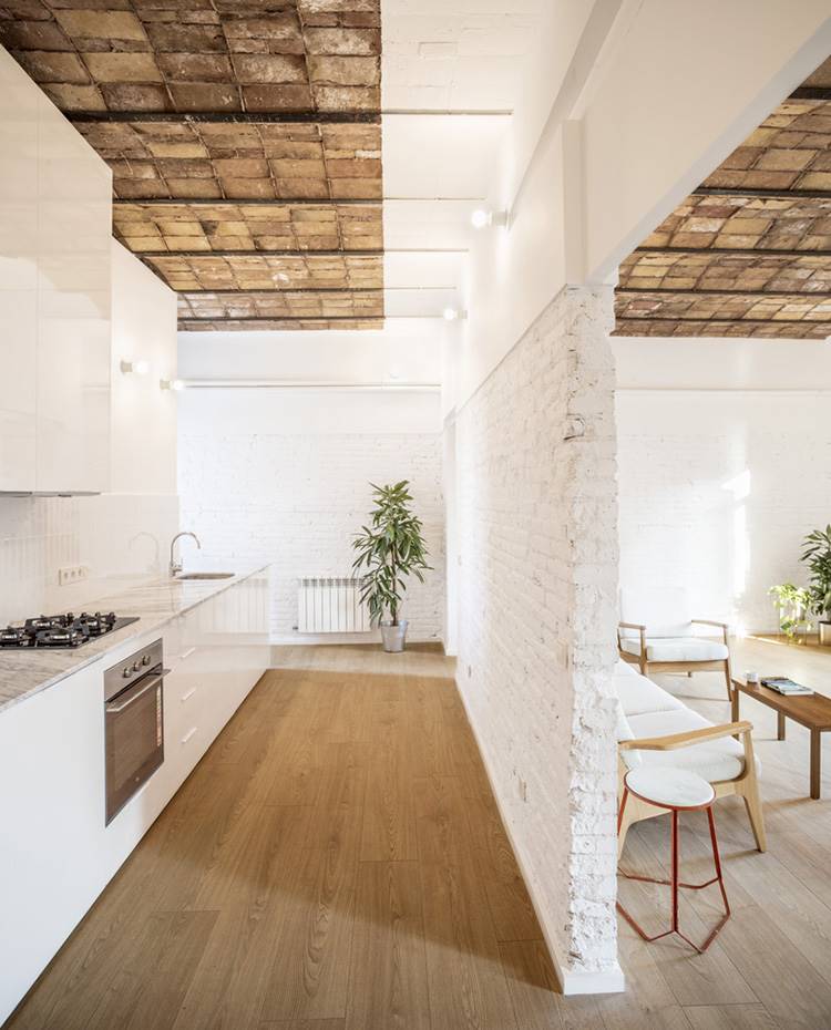 Cocina en línea, muro divisor ladrillo vista, volta catalana, zona salón. La cocina se separa visualmente del salón a través de un tabique abierto permitiendo la entrada de luz natural. La iluminación de la estancia se refuerza con luminarias tipo aplique formadas por portalámparas de PVC blanco y bombillas de LED.
