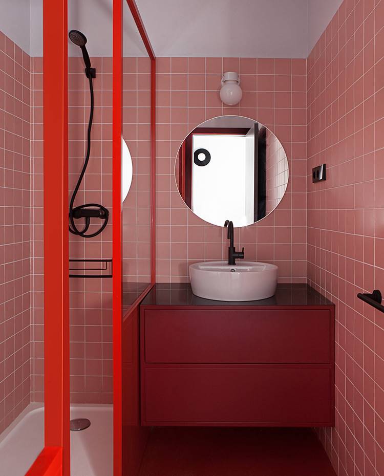 Baño con baldosas rosas, cerramiento ducha rojo, mobiliario lavabo suspendido rojo, grifería negra. En línea con el juego cromático imperante en todo el proyecto, este baño no se queda atrás y apuesta por el color rojo burdeos para teñir revestimientos, estructuras y mobiliario. 