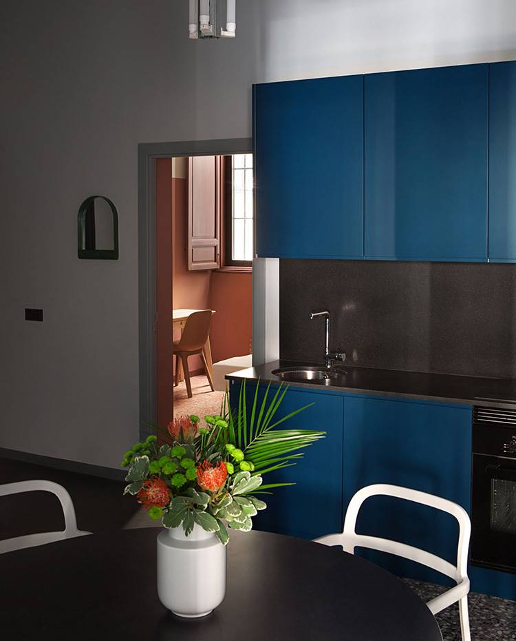 Cocina abierta a comedor, sillas blancas, jarrón con flores, cocina azul. La cocina y la sala de estar se encuentran en el centro de la vivienda y las habitaciones se extienden entorno a ellas, convirtiéndose en el punto de encuentro.