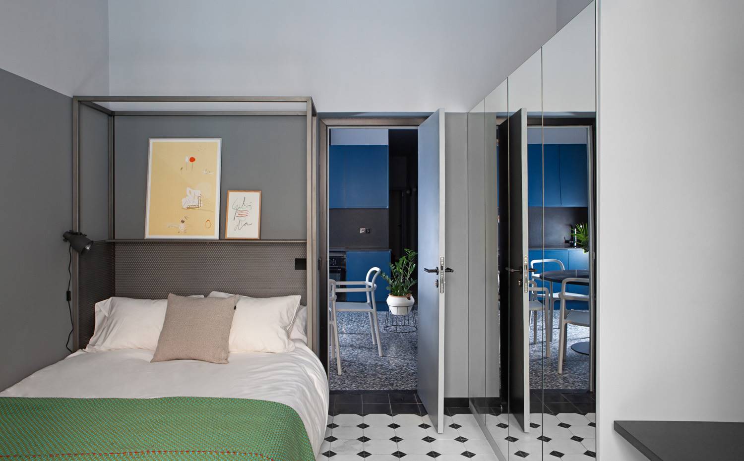 Dormitorio con estanteria de acero, lámpara aplique, armario puertas de espejo. En la habitación negra, la cama con dosel funciona como un todo en uno, acompañada de repisa, luz y cabecero. Un armario con frontal de espejo se encarga de aportar mayor sensación de amplitud a la estancia.
