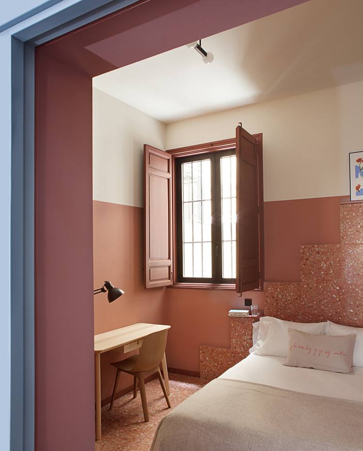 Mesa y silla zona de trabajo de madera, baldosas en suelo y paredes, marco puerta azul, cama de tonos crudos. En la habitación de la Tierra, el terrazo del suelo asciende a lo largo de la pared creando un cabecero. Su tono cálido contrasta con la frialdad del material. 