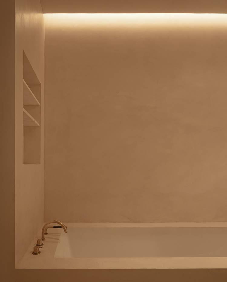 Bañera de obra, grifería encastrada, estante hueco muro perforado. El cuarto de baño se viste con grifería y apliques de acabados dorados. El equipamiento de obra mantiene el mismo criterio monocromático que el resto de la vivienda.