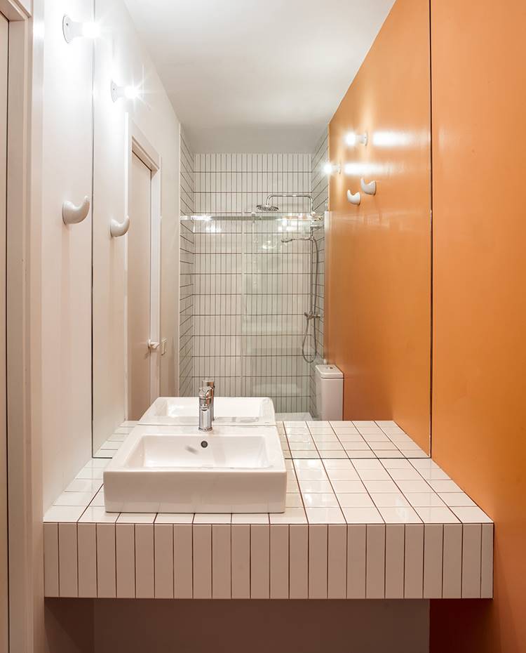 Baño, zona lavamanos, lavabo exento, revetimiento de gres blanco, pared naranja. El cuarto de baño queda oculto tras la cocina, en la zona más estrecha de la vivienda. La zona de lavabo está compuesta de lavamanos exento sobre base de obra revestida de cerámica, y se acompaña de ducha con cerramiento acristalado.
