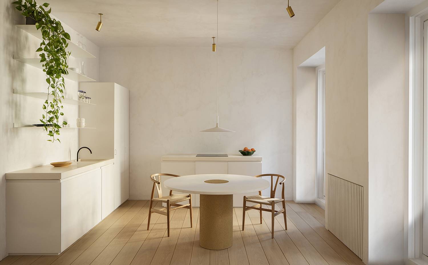 Cocina-comedor, mesas de madera, iluminación apliques techos dorados, monocromo, mesa centro redonda. La cocina, equipada con mobiliario a medida, se compone de dos frentes, uno de aguas y almacenaje y otro de cocción. El mobiliario, así como la encimera y paredes, crean un todo cromáticamente homogéneo.