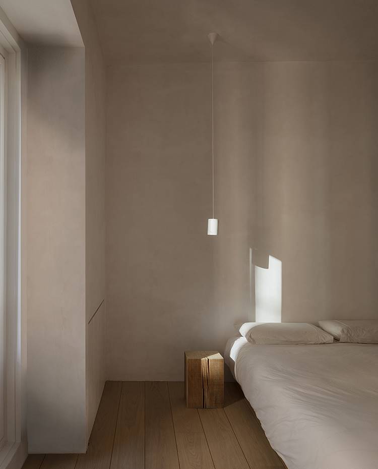 Dormitorio con cama blanca, bloque madera auxiliar, luminaria suspendida. Ajeno a elementos supérfluos, el dormitorio respira un ambiente depurado gracias a la tonalidad terrosa imperante. 