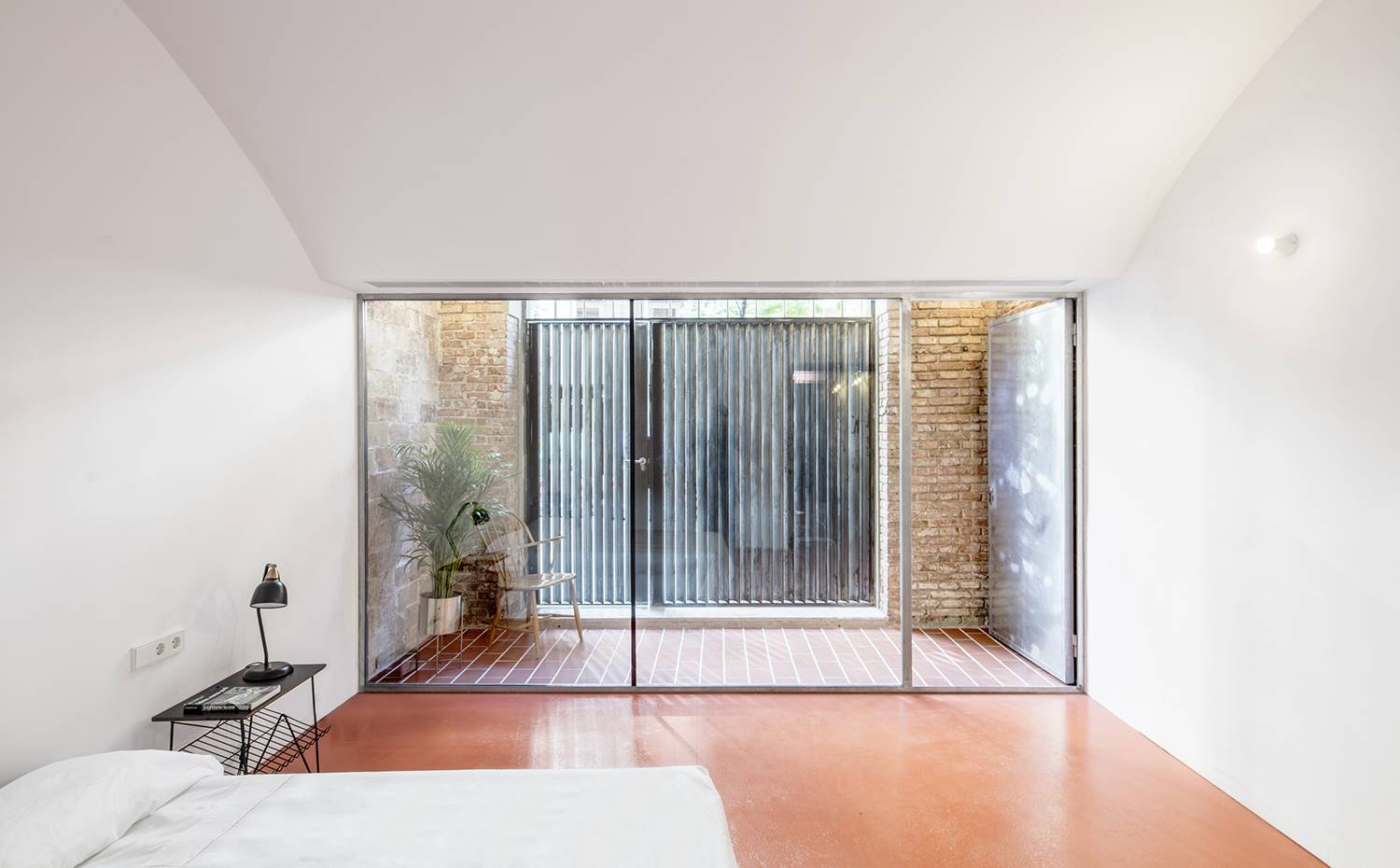 Dormitorio con cerramiento acristalado, puerta exterior al fondo, mesita auxiliar negra, suelo rojo. El dormitorio del espacio a fachada, con techo abovedado, se aísla respecto la calle a través de intercalar un patio que filtra la contaminación acústica y visual. 