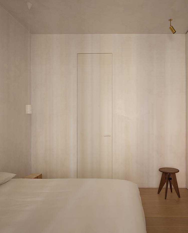 Dormitorio con paredes monocromo, taburete madera oscura. Las paredes se fusionan con los armarios de almacenaje en este dormitorio provisto de luz natural procedente de la apertura exterior a fachada. 