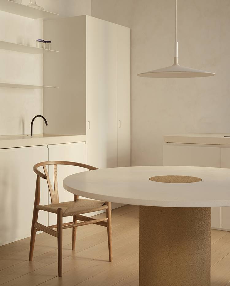Mesa comedor redonda, silla de madera, lámpara de suspensión, mobiliario cocina monocromo. Las formas curvas delimitan la zona de comedor, frente a la pureza de líneas rectas del área de cocina. La madera juega en revestimientos y mobiliario creando un ambiente cálido y sereno.