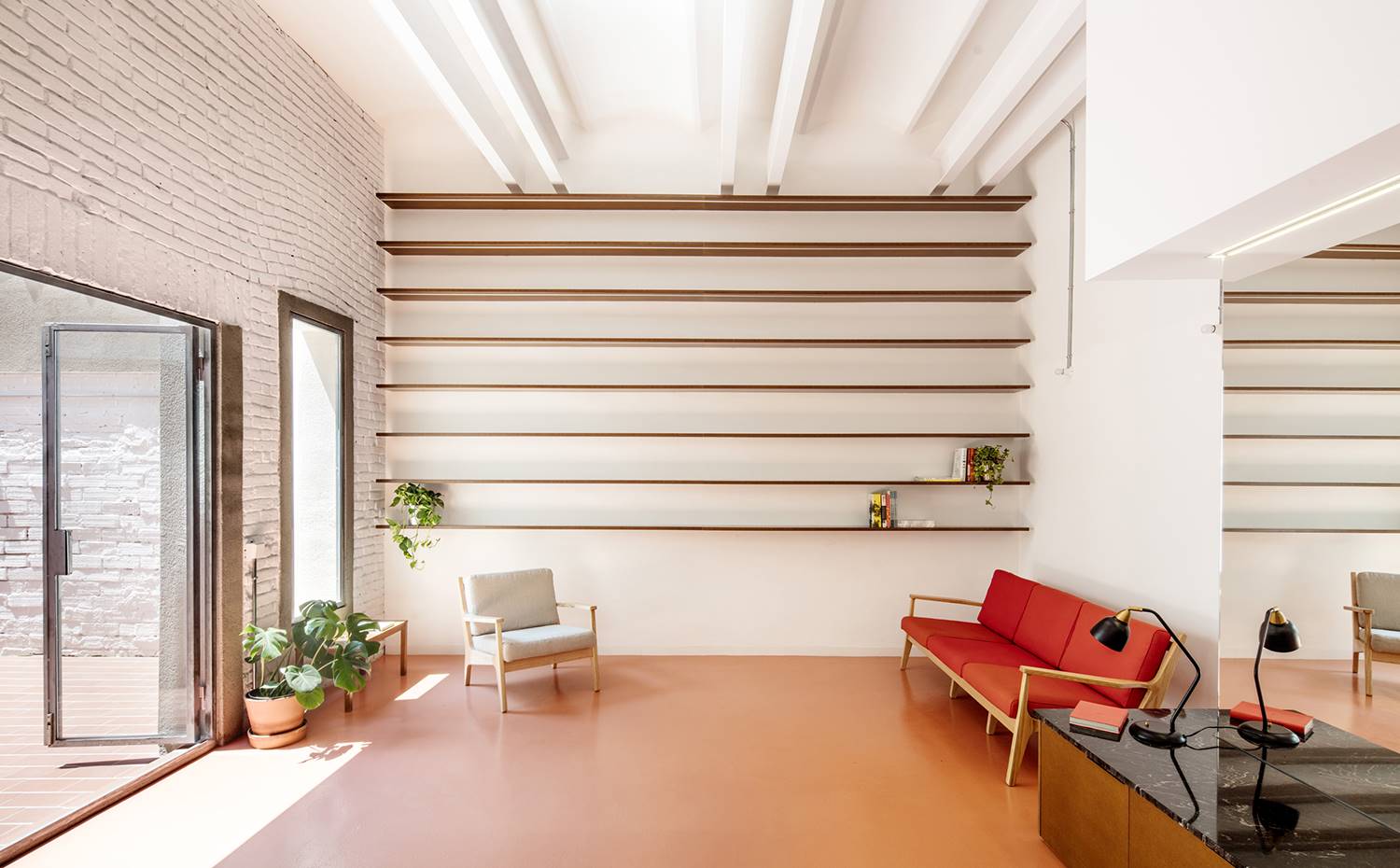 Salón con estantería de pared a pared, sofá rojo, butaca color grus claro, apertura patio exterior. La zona de día se abre hacia un patio exterior manteniendo la estética blanca de paredes y techos, y la calidez de una pátina homogénea de epoxi en color rojizo.