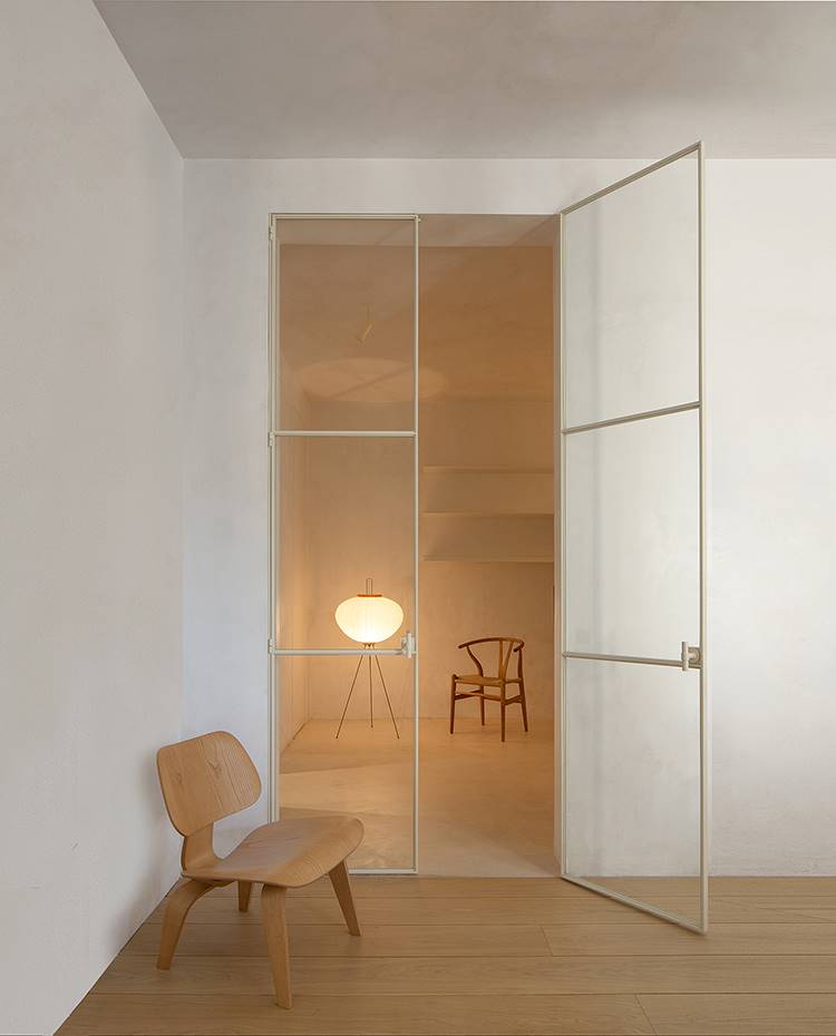 Zona de paso con butaca de madera, puerta de cristal a gran altura, iluminación de pie con silla al fondo. Presidiendo la zona de paso, el sillón Plywood Group-LCW de Vitra, en madera de fresno laminada y moldeada, frente a una puerta de cristal transparente de grandes dimensiones.