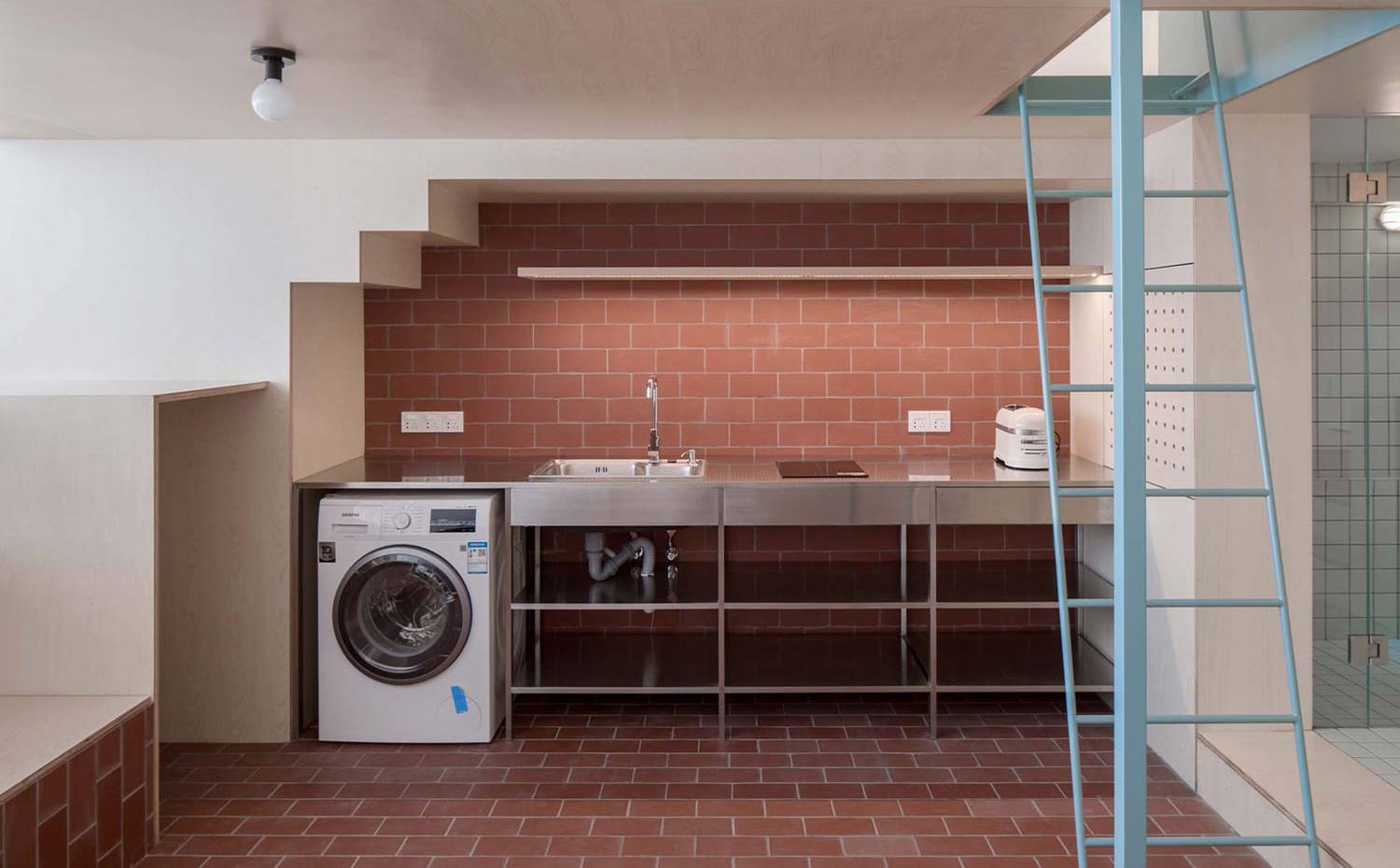 Cocina frontal, revestida de baldosa roja, escaleras hacia la zona de estudio. La cocina, de aspecto industrial aunque muy mínimal se situa bajo el hueco de la escalera que accede a la planta superior.