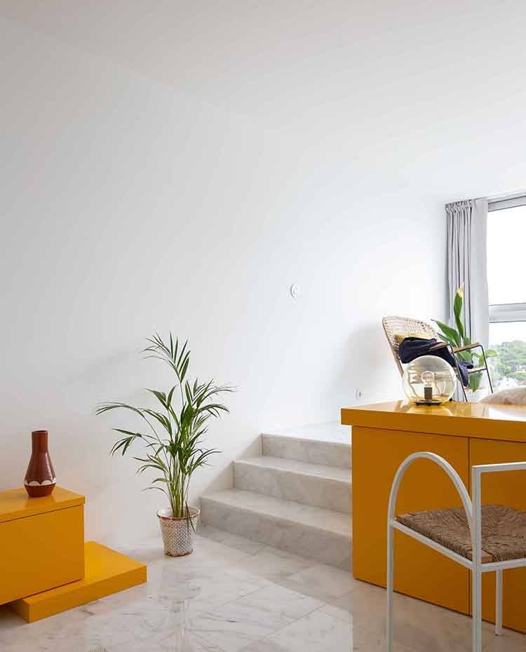 Detalle escalera a zona dormitorio, mobiliario lacado en amarillo, silla blanca con asiento de fibra. La zona del dormitorio queda elevada, accediendo a ella a través de dos tramos de escaleras situadas en los laterales de la planta. Queda flanqueada por el segundo de los tres muebles amarillos que visten el lugar. 