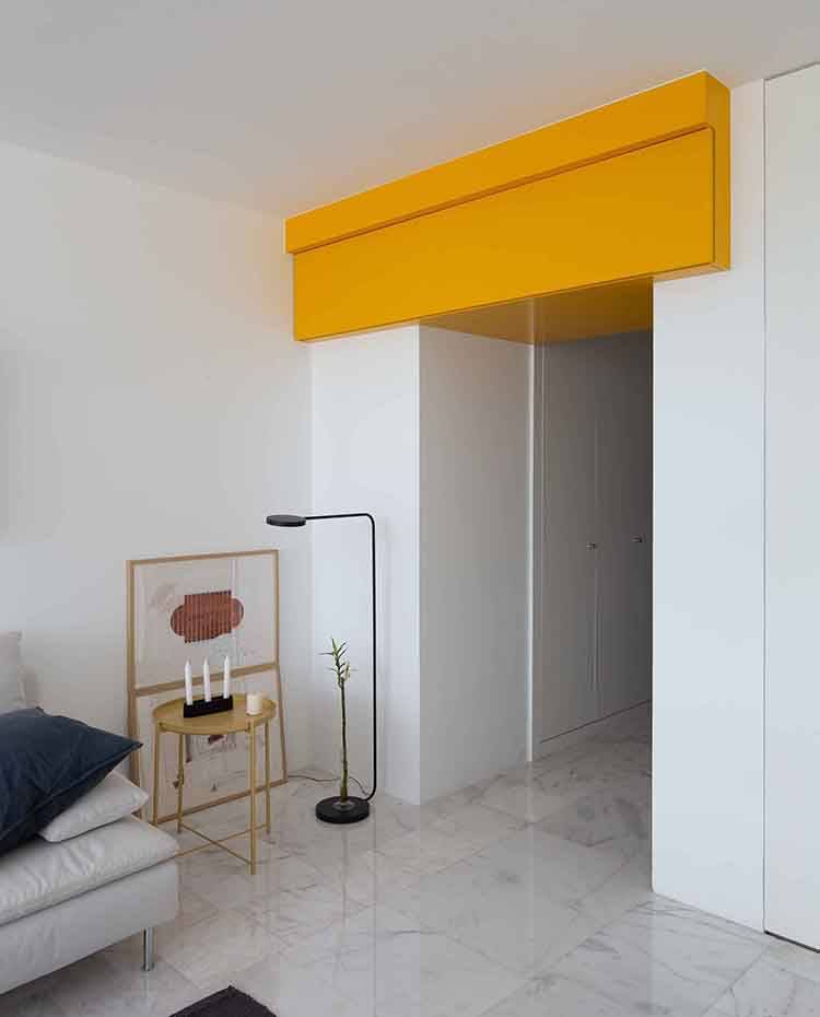 Zona de paso junto a salón, mueble en bajotecho, lacado amarillo, mesita auxiliar metalizada, luminaria de pie tubular. El tercero de los elementos que conforman la composición interior del espacio es este armario a gran altura que aprovecha el interior del bajo techo y lo destina a almacenamiento. 