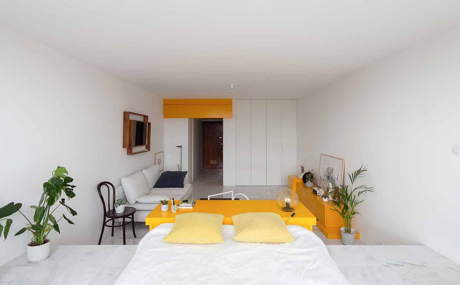 Zona dormitorio, mueble contenedor amarillo separador de ambiente, silla Thonet, sofrá blanco,  mueble bajo amarillo. El apartamento tipo estudio se proyecta como una caja en blanco, cediendo el protagonismo (con permiso de las panorámica) a las tres piezas de mobiliario de color amarillo.  