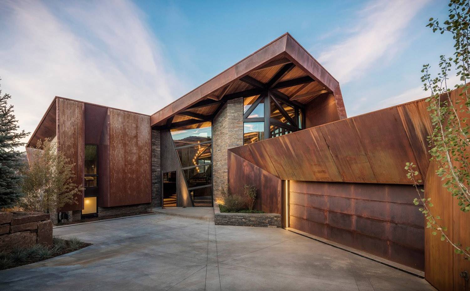Estructura exterior vivienda de acero corten y miedra, entrada con cerramientos de cristal y madera.. La robusta estructura de acero de la casa imita la topografía del área circundante, mejorando las conexiones visuales con el paisaje y al mismo tiempo dibujándola hacia adentro.