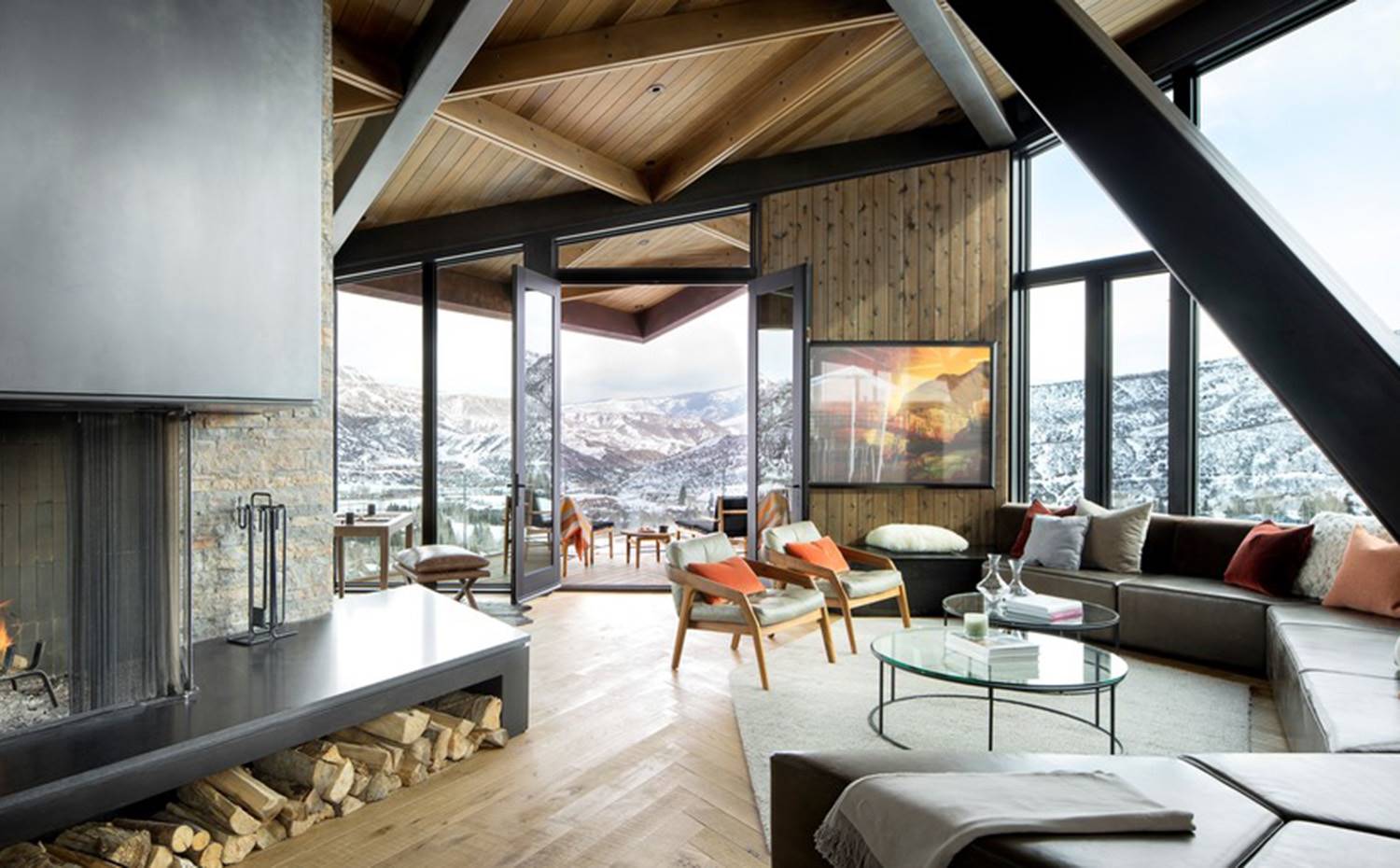Salón con amplio sofá modular, dos mesas auxiliares de cristal y acero, dos butacas de madera, amplia chimenea abierta en pared de piedra, revestimiento de madera, exterior nevado.. Las paredes de vidrio del piso al techo en las áreas comunes forman la punta del plano triangular y ofrecen amplias vistas del paisaje circundante. 