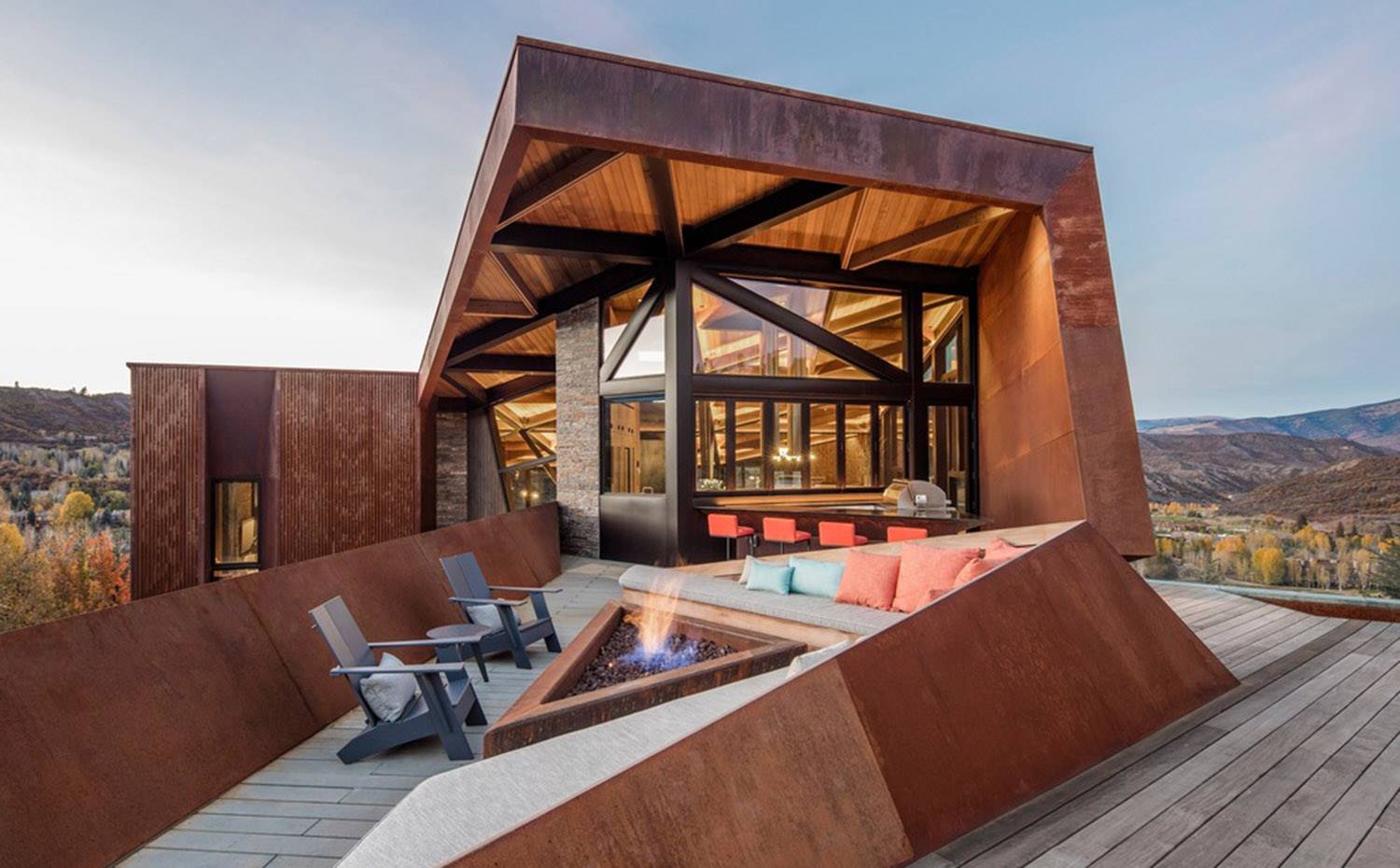 Terraza exterior con zona de estar en sofá corrido triangular, hamacas grises, chimenea central a suelo. Este retiro de montaña se diseñó en torno a la idea de que un lugar puede profundizar las conexiones entre las personas, las familias y el paisaje que aman.