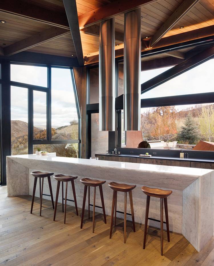 Zona de cocina provista de isla, con office y taburetes de madera. Dos campanas extractoras tubulares de hacero, cubierta de madera. La amplia zona de cocina está provista con dos frentes de trabajo, uno de ellos integrado en una gran isla, con taburetes de madera frente a un área destinada a office.