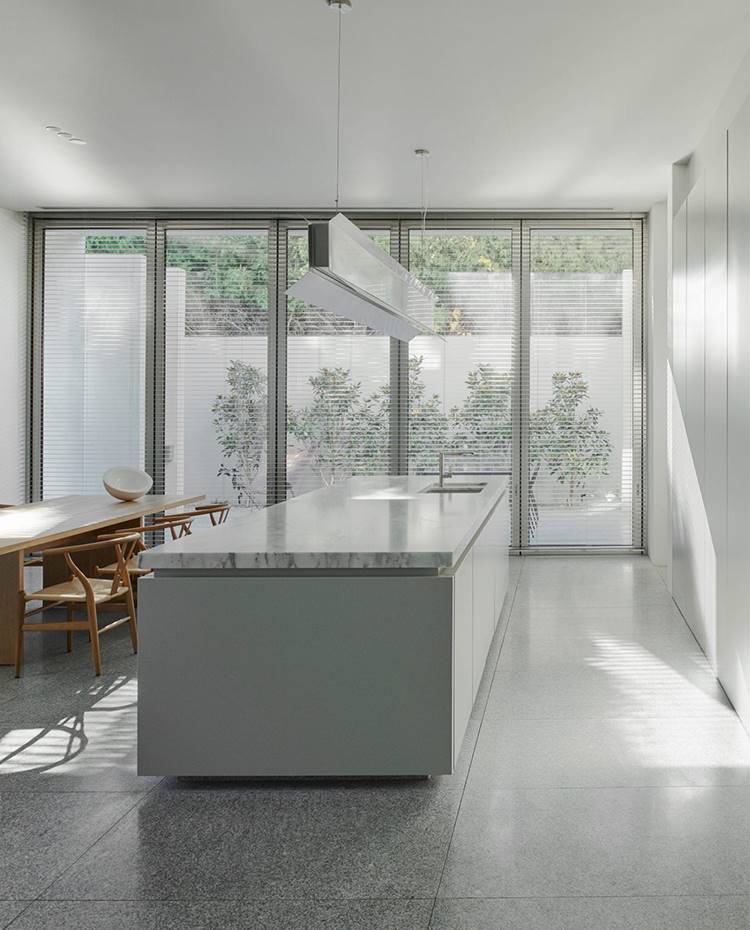 Isla de cocina, mobiliario gris, sobre encimera mármol, fregadero, lámpara de suspensión metalizada, mesa de comedor de madera. Parte de la cocina con isla se oculta tras cerramientos laminados. Sobre la encimera de trabajo, una lámpara suspendida de diseño personalizado que ilumina directamente sobre el área y arroja luz difusa hacia el techo. 