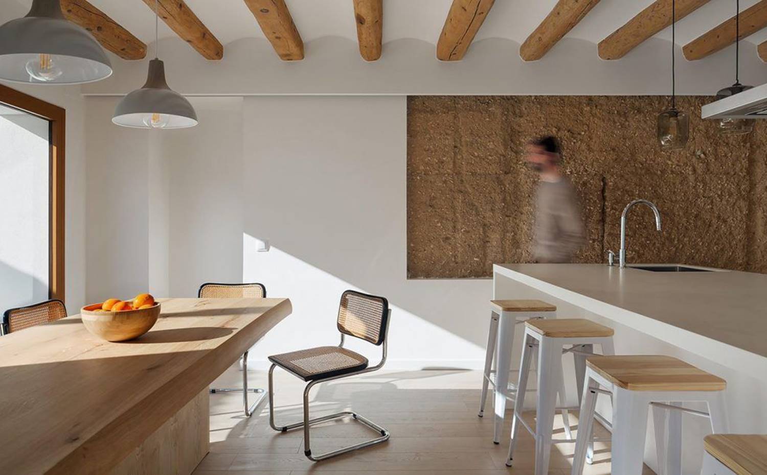 Cocina con mesa de office de madera, silla de fibra y estructura modular, taburetes blancos junto a isla de trabajo, techo con vigas de madera. El equilibrio entre el legado histórico de la casa y las soluciones funcionales y vanguardistas se materializa a lo largo de todo el proyecto, como la cocina.