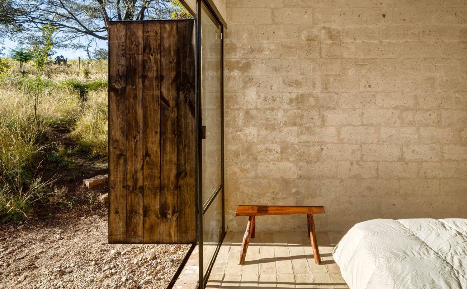 Detalle interior dormitorio, cerramiento cristal con frente de madera, banco de madera, cama con plumón, exterior naturaleza y tierra. El estudio de arquitectos PPAA eligió arcilla para los suelos de la casa y madera local para otros elementos de construcción. 