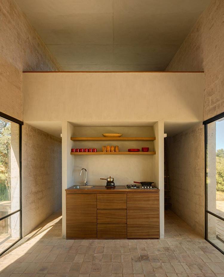 Espacio a doble altura con cocina en hueco de hormigón, armarios bajos de madera, estantes superiores de madera, con altillo superior. Suelos de gresite, revestimientos de hormigón. Este extremo de la casa a doble altura integra la zona de cocina, tras la cual se ubica el baño, y un dormitorio en la parte superior. 