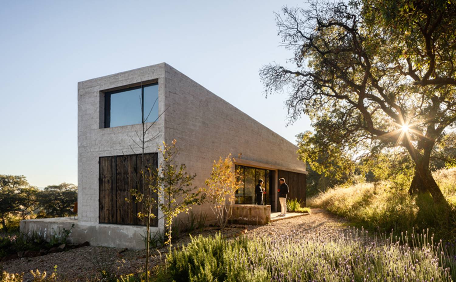 Exterior vivienda, Cerramiento madera y cristal, entorno natural, terraza de hormigón. La vivienda presenta un volumen de piedra de forma rectangular, con un perfil de techo inclinado que se asienta a un entresuelo en uno de sus extremos. 