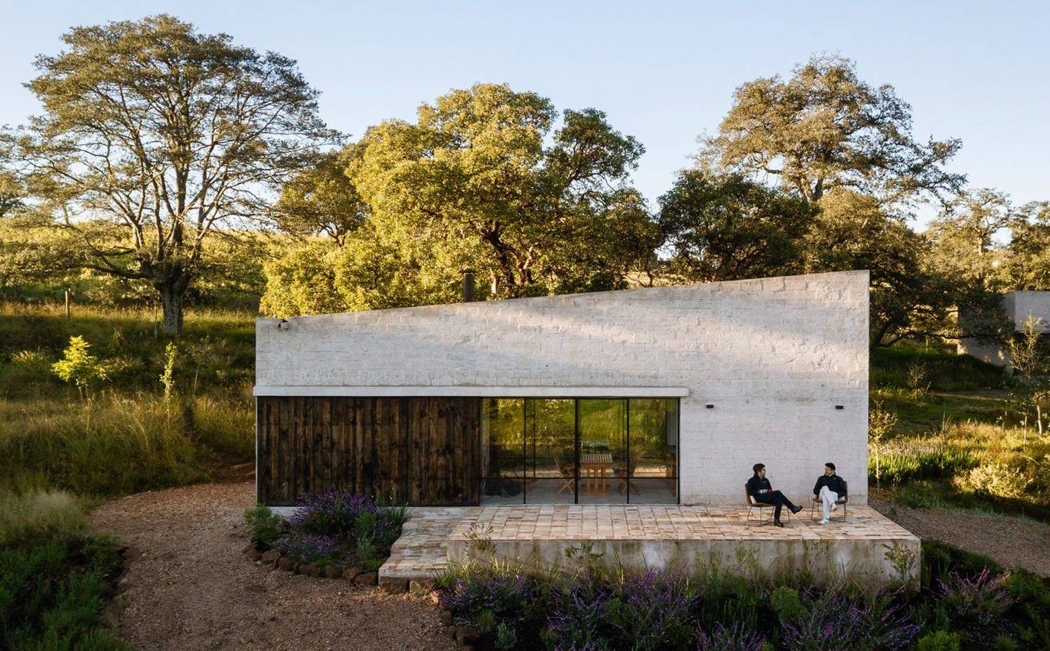 Exterior vivienda, terraza con dos personas en sillas, entorno arbolado. Los propietarios, dos hermanos amantes de los deportes de aventura, encontraron por casualidad la parcela a las afueras de la ciudad de Aculco, al noreste de Ciudad de México, durante un viaje de escalada. 