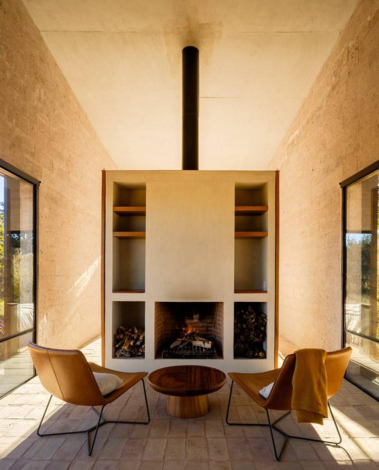 Mueble a medida con chimenea y estanterías, mesa de centro redonda, butacas de piel con estructura turbular, paredes hormigón, cerramientos cristal. La búsqueda por la simplicidad, la mínima necesidad de mantenimiento y el diálogo íntimo con el entorno ha dado como resultado un proyecto sobrio, con módulos de obra, como este que integra la chimenea, que actúan como separadores de espacios.